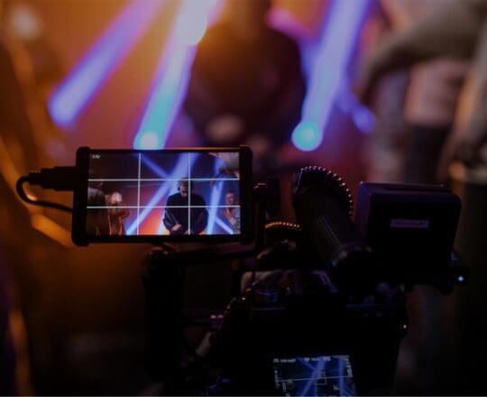 Broadcast camera filming a live show with a focus on the camera's monitor displaying the scene.