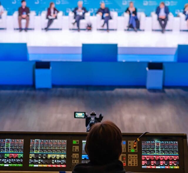 Camera operator controlling equipment while filming a live event panel discussion.
