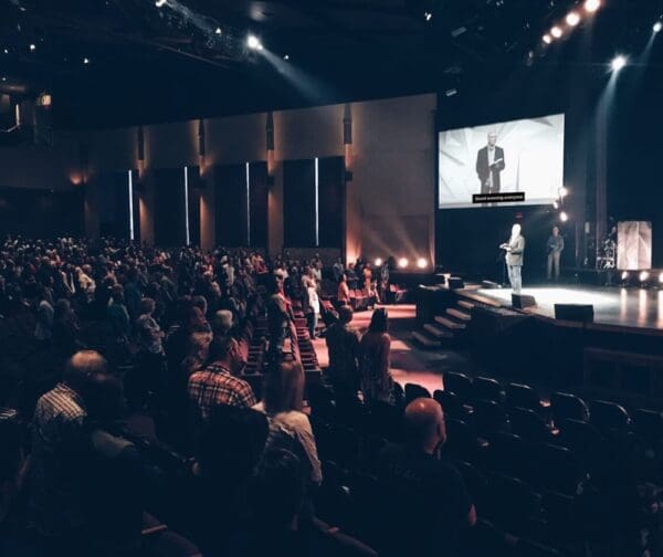 Audience standing at a live event with a speaker on stage, captions displayed for accessibility.