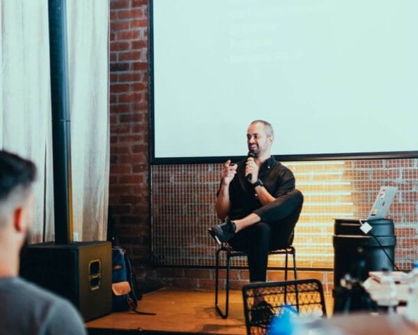 Speaker addressing an audience during a presentation with a projector screen in the background.