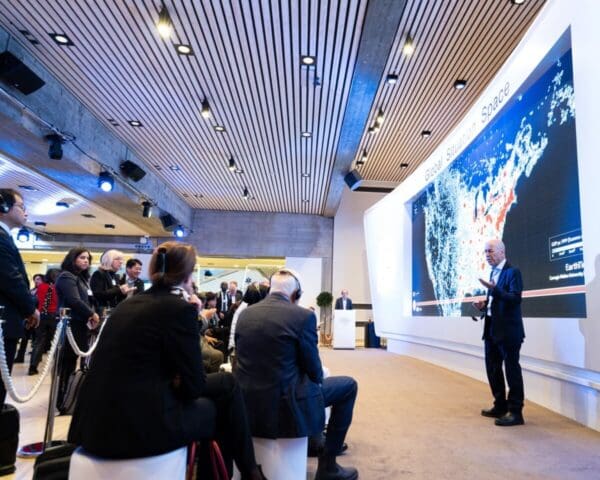 Speaker presenting at a conference with a large screen showing a global map and an audience listening.