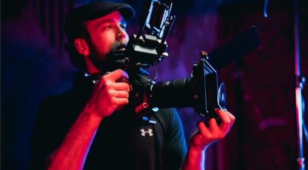 Person holding a broadcast camera with a lens and monitor in a studio setting.
