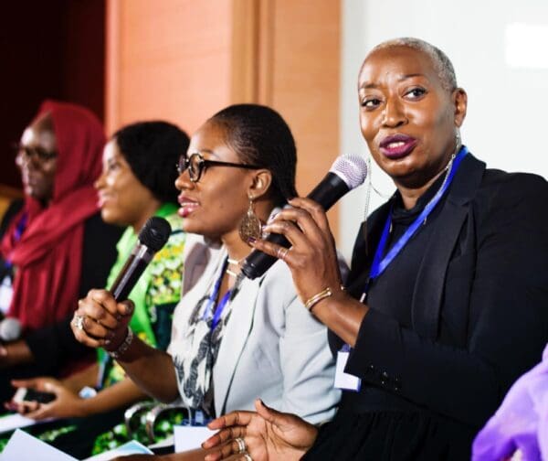 Group of multicultural women speaking on stage with microphones during a public speaking event.